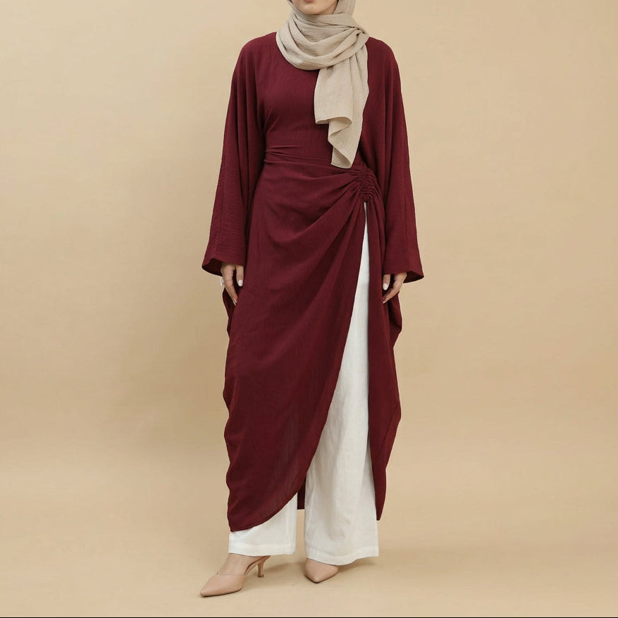 A model wearing a long-sleeved, deep burgundy wrap-style tunic with a twisted waist detail, paired with white wide-leg pants, against a plain white studio background.
