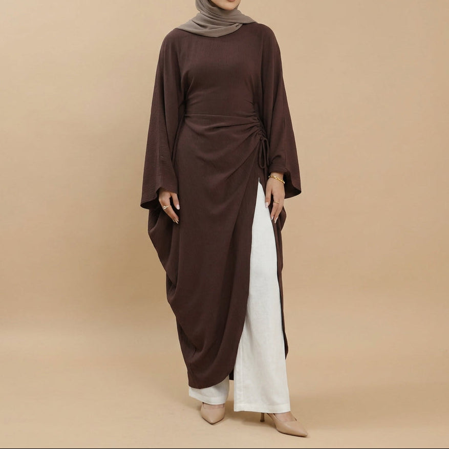 A model wearing a dark brown long-sleeved maxi dress with draped, asymmetrical layers and white wide-leg pants, photographed against a seamless white studio background.