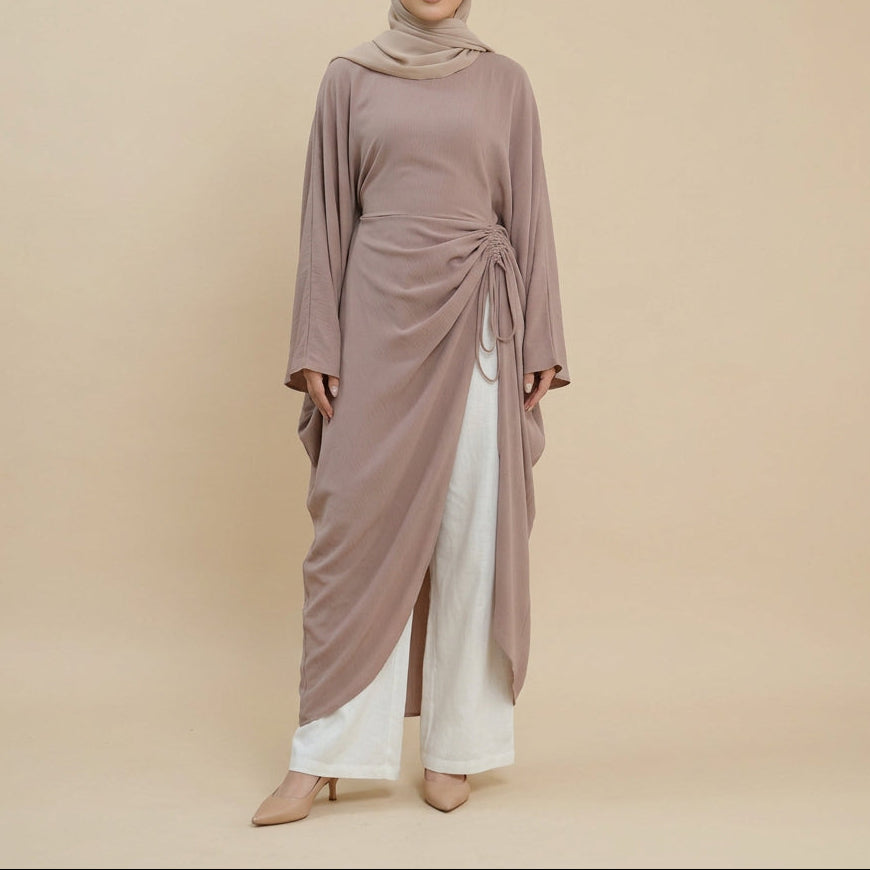 A model wearing a loose, draped taupe-brown long-sleeve tunic top over white wide-leg pants, standing against a plain white studio background.