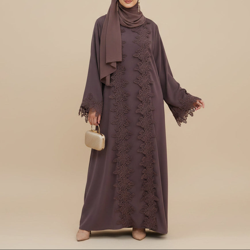 A woman models a long-sleeved, floor-length modest dress in a muted plum-brown color with matching floral lace panels down the front and a coordinating hijab. She is holding a small beige clutch against a solid beige background.