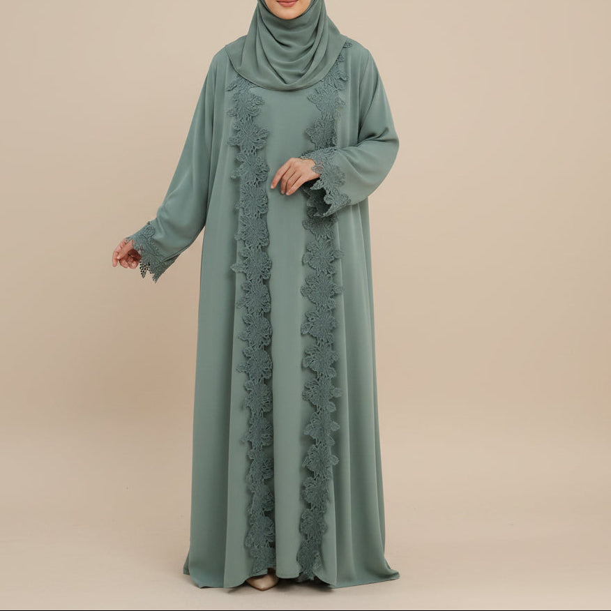 A woman modeling a sage green modest outfit consisting of a long abaya with lace trim and a matching headscarf, standing against a light gray studio background.