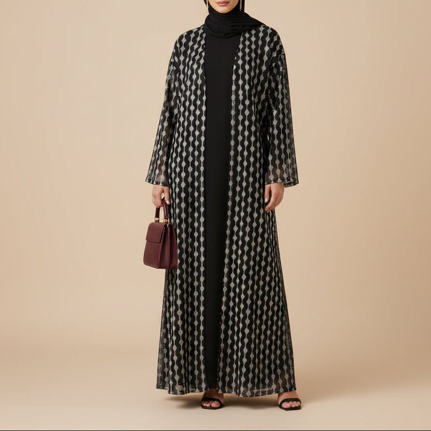 A woman modeling a long black abaya with a white geometric pattern, worn over a black inner dress and black hijab, holding a beige clutch against a plain off-white studio background.