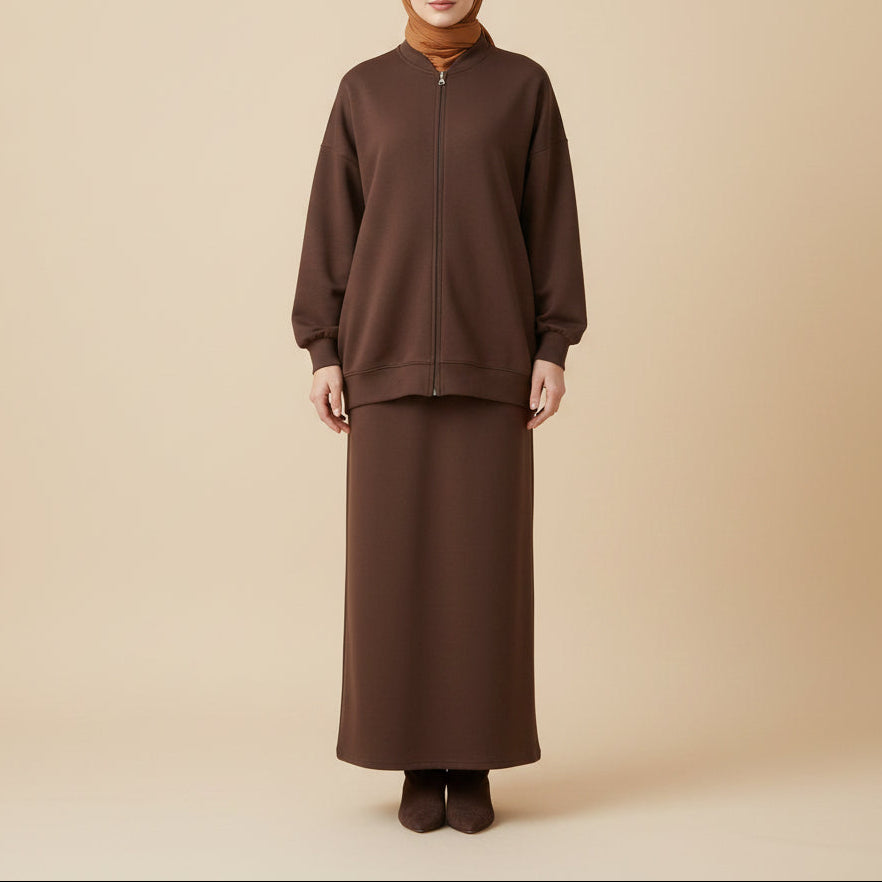 A dark brown two-piece outfit displayed on a mannequin, featuring a cropped zip-up jacket and a matching long skirt, set against a white background with decorative pampas grass and a white armchair.