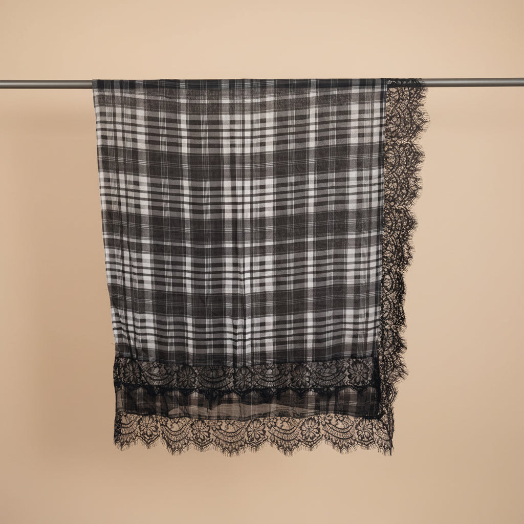 A black and white plaid scarf with intricate lace trim on all three edges, hanging from a metal rod against a plain white wall.