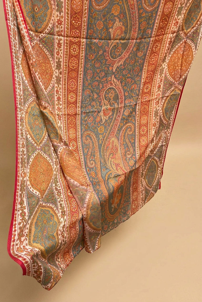 A hand holding up a patterned fabric, likely a sari, featuring a detailed paisley and floral design in orange, gold, blue, and red with a red border.