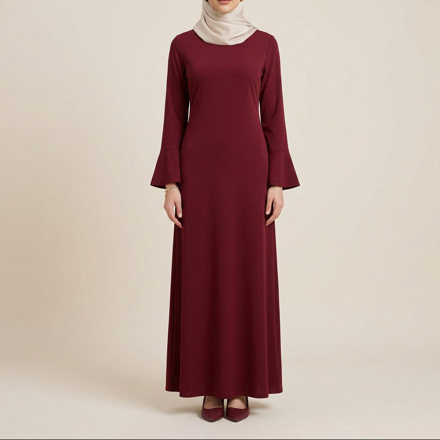 A long-sleeve, floor-length maroon dress displayed on a mannequin against a white paneled wall with a green palm leaf in the corner.