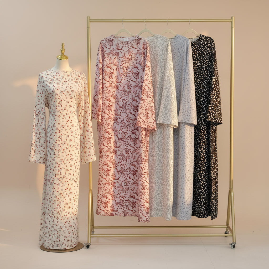 Four long-sleeve floral maxi dresses displayed on a gold clothing rack, with a matching dress on a mannequin to the left. The dresses feature different floral patterns and colors: pink and red on cream, light pink and white, light blue and white, and black and white.