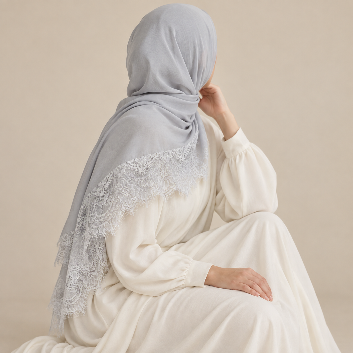 A woman wearing a light gray hijab with white lace trim, styled over an off-white long-sleeve dress, shown from the back against a warm beige background.