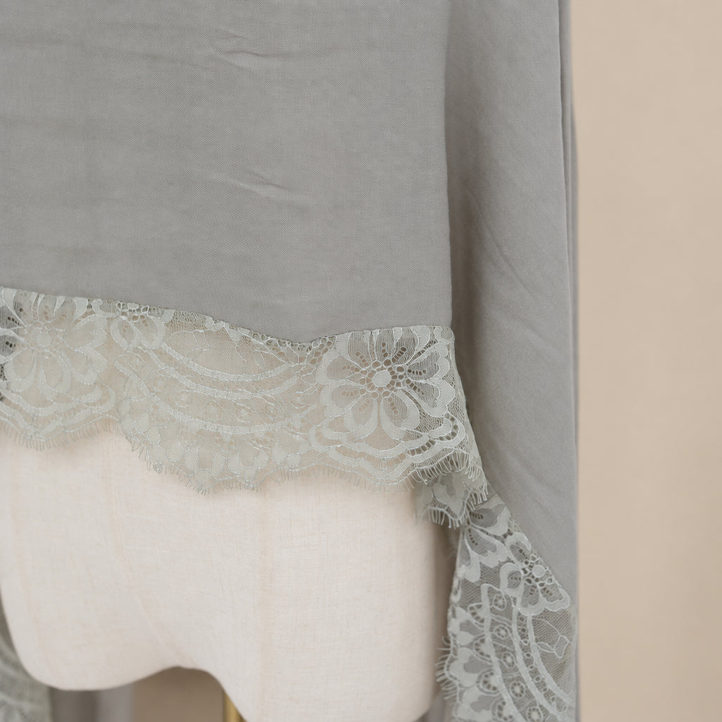 A close-up of a light taupe-gray fabric draped over a cream-colored dress form, featuring a decorative white floral lace trim along the bottom edge.