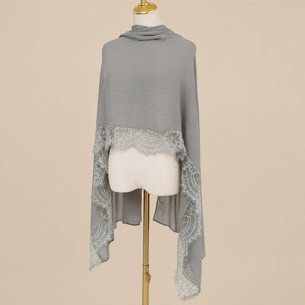 A taupe gray shawl with a scalloped lace hem, draped on a cream dress form mannequin with a gold stand, set against a white background with pampas grass on the left.