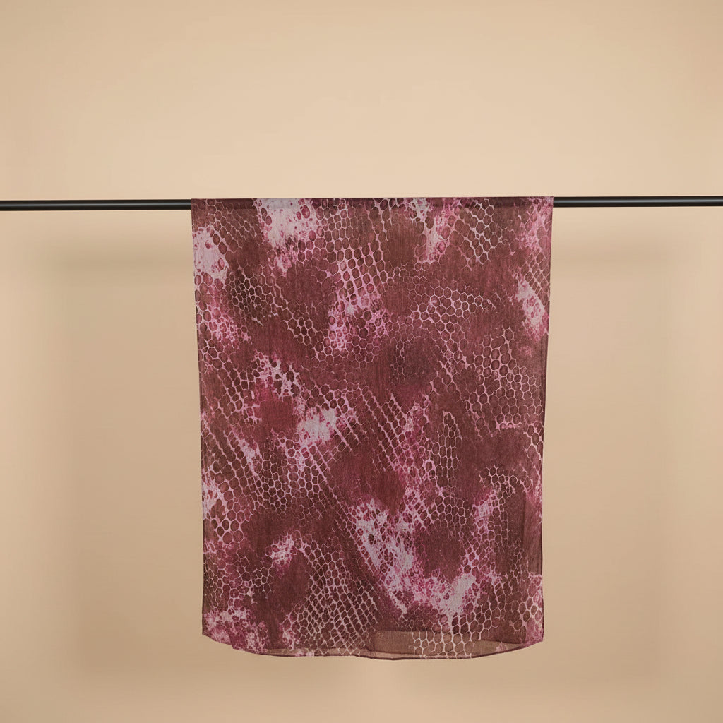 A sheer scarf with a snakeskin pattern in shades of burgundy, pink, and brown, hanging from a black clothing rack against a plain white wall.