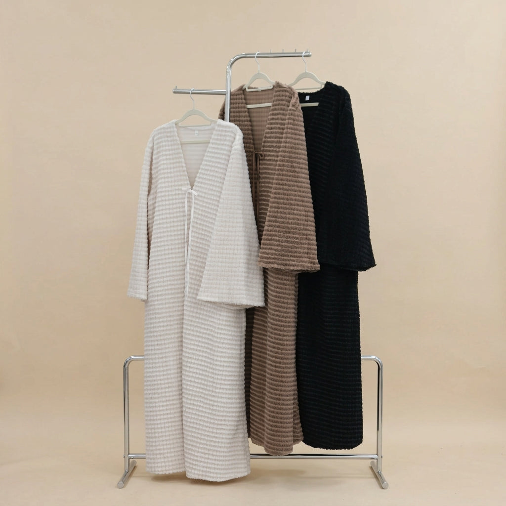 Three textured waffle-knit long cardigans in cream, taupe, and black hanging on a metal rack against a white paneled wall with a green plant in the background.