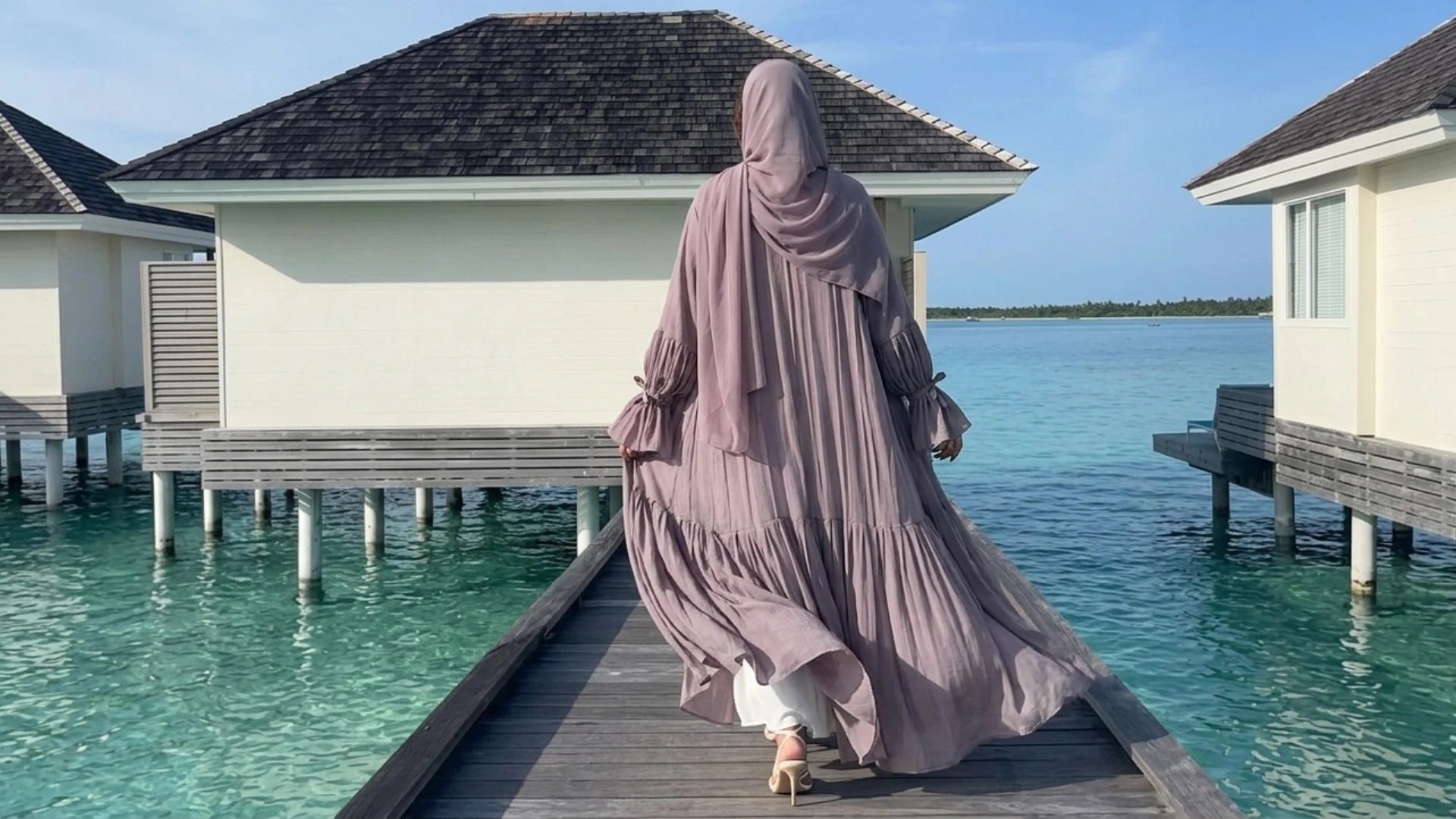 Woman wearing a flowing modest hijab abaya in soft mauve walking on a luxury ocean resort boardwalk, elegant Islamic modest fashion for women by Mdoest