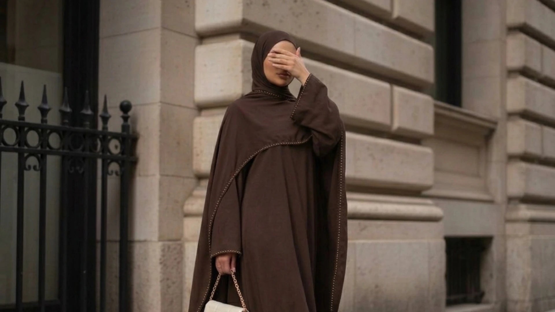 A model wearing a deep chocolate brown modest abaya with gold beaded trim details and a matching hijab by Try Modest, holding a cream handbag in an urban city setting