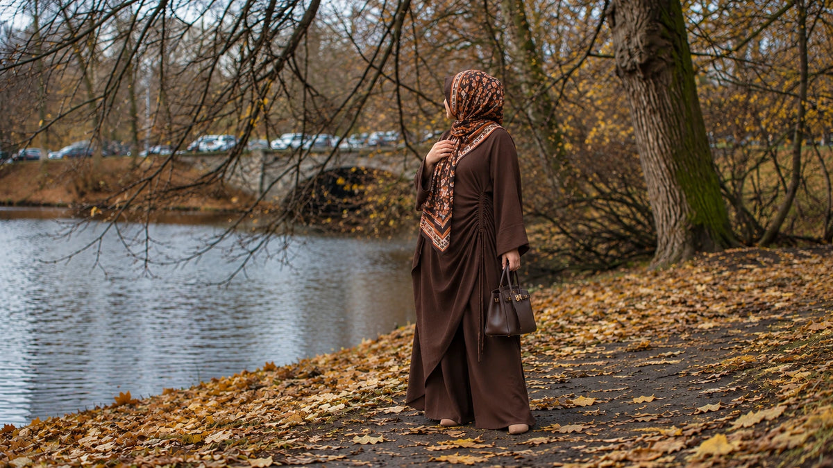Try Modest Estelle Ruched Side-Tie Abaya Co-Ord Set in coffee brown, styled with a printed hijab beside an autumn lakeside with golden fallen leaves