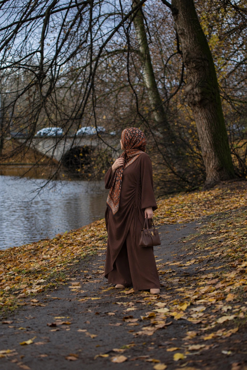 Try Modest Estelle Ruched Side-Tie Abaya Co-Ord Set in coffee brown, styled with a printed hijab beside an autumn lakeside with golden fallen leaves