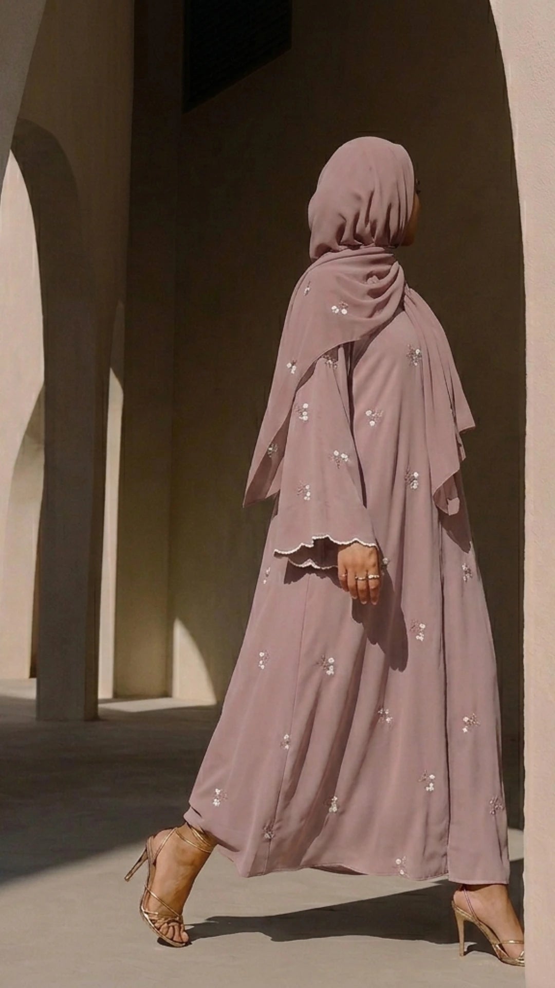 A woman walking in a dusty pink mauve embroidered abaya and matching hijab by Try Modest, paired with gold high heels against an architectural archway backdrop.