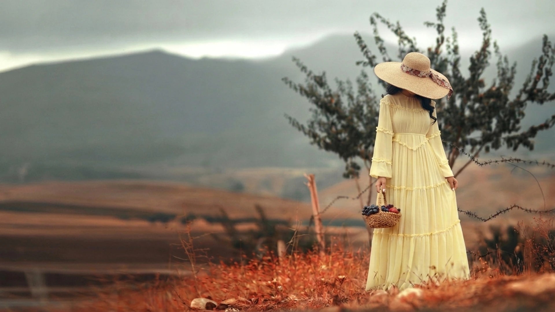 Woman in a pastel yellow summer abaya dress and wide-brim straw hat holding a fruit basket in a scenic outdoor field, by Try Modest fashion brand.