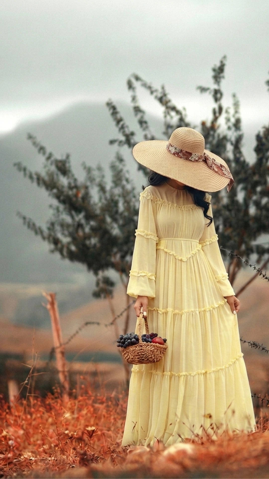 Woman in a pastel yellow summer abaya dress and wide-brim straw hat holding a fruit basket in a scenic outdoor field, by Try Modest fashion brand.